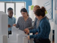 Diverse group of colleagues working on building model design with blueprints plan. Coworkers using maquette and industrial sketch to create architecture project, brainstorming ideas.